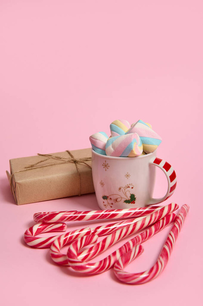 Kávový hrnek s barevnými marshmallows, vánoční dárek v řemeslné balení papíru a pruhované cukrové tyčinky na růžové pastelové pozadí. Vánoční a novoroční koncept s kopírovacím prostorem pro reklamu - Fotografie, Obrázek