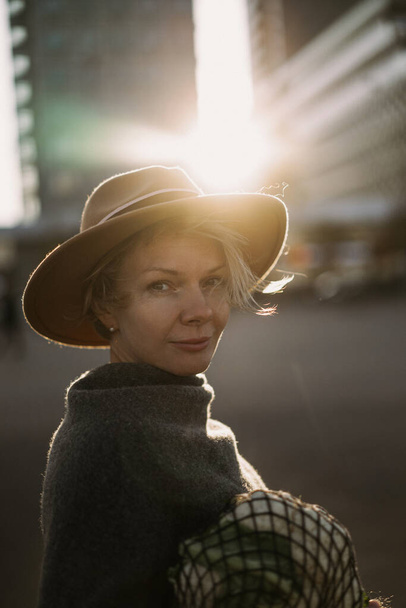 Beautiful middle-aged woman in a hat with a short haircut in the center of a big city. Close-up portrait, soft backlight. - Fotografie, Obrázek