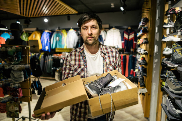 Blanke mannelijke verkoper consultant in outdoor winkel presenteren wandelschoenen aan koper. Een verkoper in een sportwinkel helpt een shopper bij het kiezen van wandelschoenen. Verkoper in sportwinkel. - Foto, afbeelding