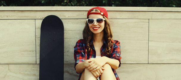 Happy smiling young woman sitting with skateboard wearing a baseball cap on city street - Foto, immagini