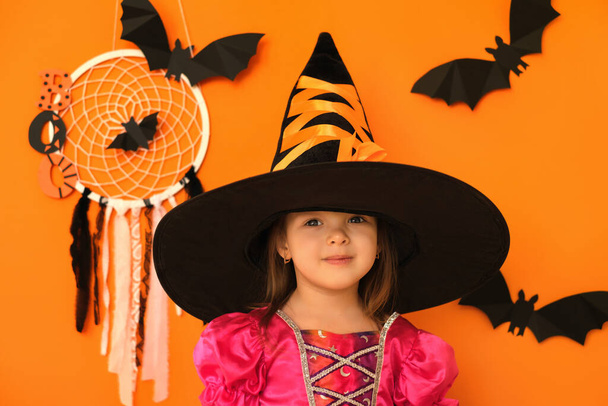 Jolie petite fille souriante positive sur fond de studio orange avec décor d'Halloween. Une fille avec un grand chapeau noir et une robe de sorcière. Dream Catcher avec toiles d'araignée, rubans et chauves-souris - Photo, image