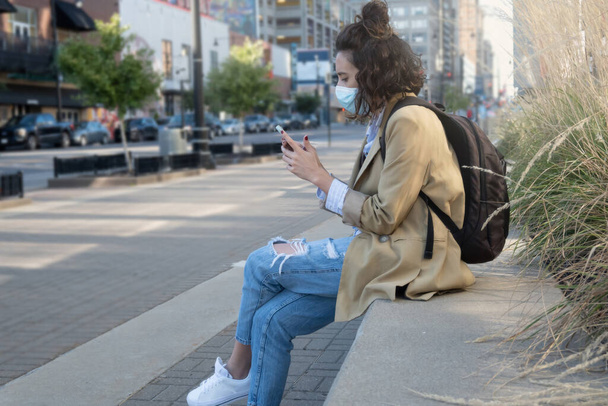 Tiro frontal horizontal de mujer sentada con máscara enviando mensaje con teléfono móvil y mochila colgante, con espacio para copiar. - Foto, Imagen