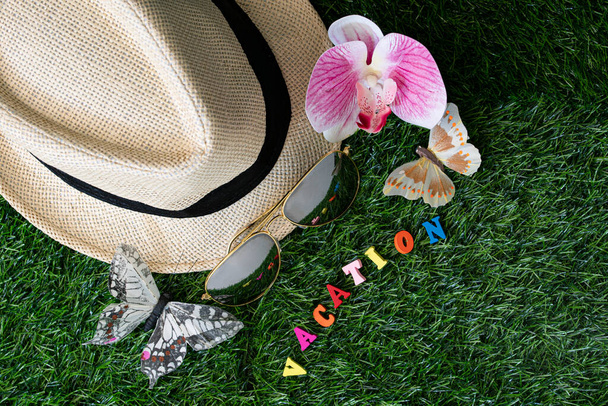 Concepto de vacaciones. Diseño de sombrero de paja, mariposa falsa, orquídea, gafas de sol y alfabeto de madera con texto VACACIÓN. - Foto, Imagen