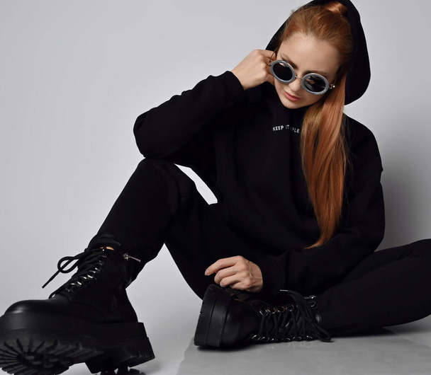 Red-haired woman in trendy black sportswear sits on floor with her cowl on her head looking at her massive army shoes - 写真・画像