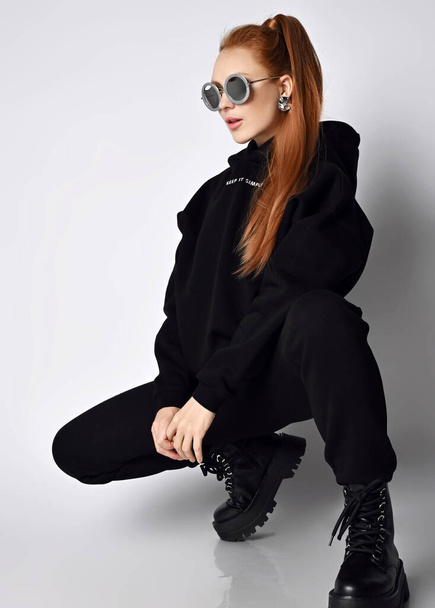 Slim fashionable redhead woman in trendy black sportswear hoodie, pants and massive brutal shoes sits squatted sideways - Fotografie, Obrázek
