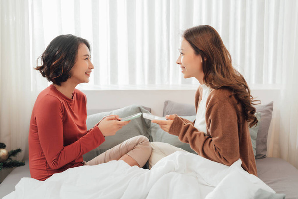 Lächelnd ruhen sich zwei junge asiatische Paare auf einem weißen Bett aus und spielen fröhlich mit Smartphone und sozialen Medien. - Foto, Bild