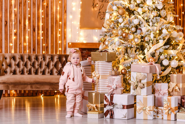 cute smiling baby girl in pink hoodie and joggers stands near the Christmas tree with gifts. - Foto, Imagen