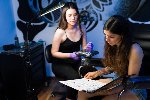 Mooie jonge vrouw kijkend naar de schetsen in de tattoo salon met een professionele tattoo artiest  - Foto, afbeelding