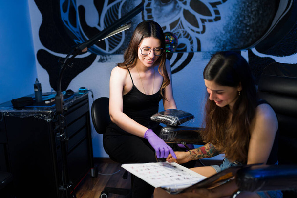 Tetszik ez a tetoválás. Boldog profi művész mutatja tetoválás vázlatok egy fiatal nő a stúdióban  - Fotó, kép