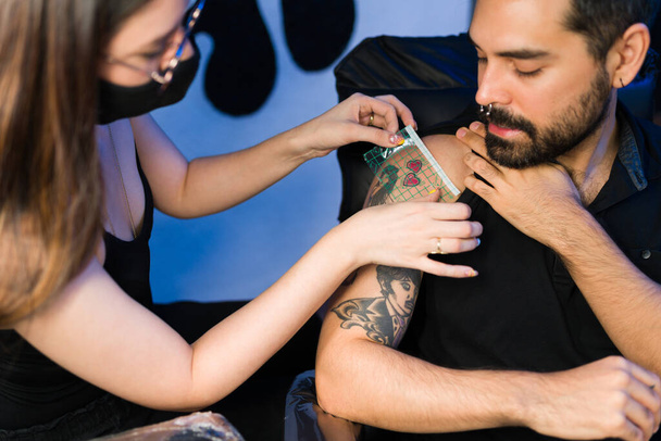 Joven artista que mide un pequeño tatuaje en el brazo de un cliente masculino en su salón de tatuajes - Foto, Imagen