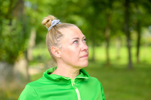 Nachdenkliche blonde Frau mittleren Alters mit zu einem Knoten gebundenen Haaren auf dem Kopf, die mit nachdenklichem Gesichtsausdruck in einem bewaldeten Park aufsteht - Foto, Bild