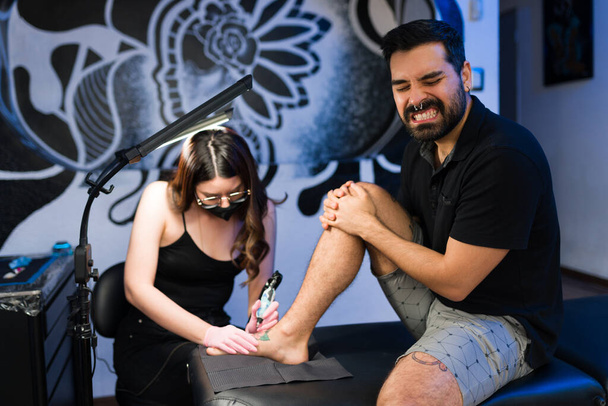 Sconvolto giovane uomo in un sacco di dolore sentirsi male e la sofferenza, mentre ottenere un nuovo tatuaggio sul piede in studio - Foto, immagini