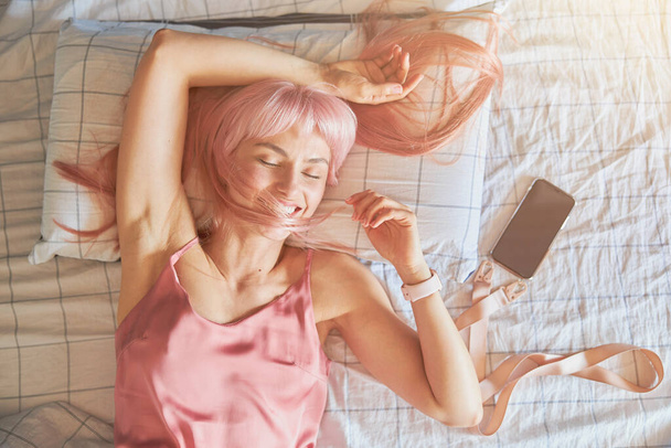 Relajada mujer sostiene el mechón de pelo rosa por encima de la cara acostado cerca del teléfono en la cama - Foto, Imagen
