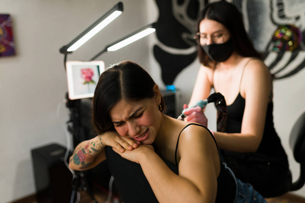 Es tut sehr weh. Gestresste junge Frau lässt sich im Tintenstudio von einer professionellen Tätowiererin tätowieren - Foto, Bild