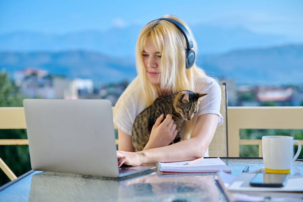 Female student studies at home online using laptop. Teenager sitting on outdoor balcony with pet cat in headphones, school notebooks, looking at screen. E-learning, modern technologies in education - Fotoğraf, Görsel