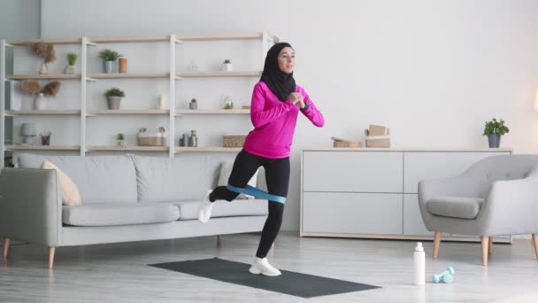 Mujer musulmana joven positiva haciendo ejercicio con goma elástica, haciendo práctica deportiva para piernas con carga extra en casa - Metraje, vídeo