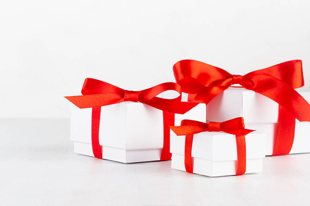 Three white paper gift boxes with red ribbon and bow in soft light interior on white table, copy space, closeup. - Foto, Imagem