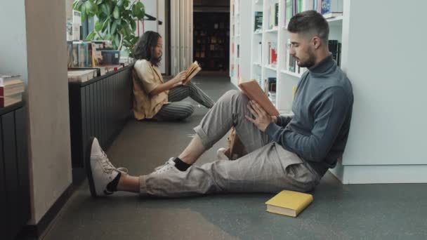Steek slowmo shot van jonge multi-etnische man en vrouw lezen terwijl zitten op de vloer naast boekenkast in de moderne bibliotheek - Video