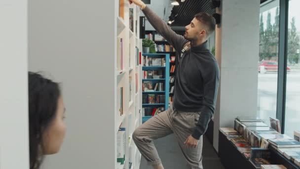 Közepes lassítású felvétel fiatal Vegyes fajú férfi egyetemi hallgató hátizsák nyúlik könyvet könyvespolcon és elkezd olvasni, miközben időt tölt a modern könyvtárban - Felvétel, videó