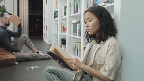 Medium slowmo schot van vrouwelijke Aziatische universiteit student zitten op de vloer van hedendaagse bibliotheek leesboek - Video