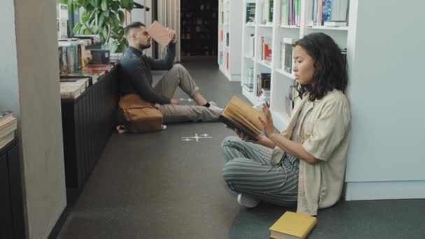 Dollying-in slowmo van twee jonge multi-etnische mannelijke en vrouwelijke universiteitsstudenten die boeken lezen terwijl ze in de moderne bibliotheek op de vloer zitten - Video