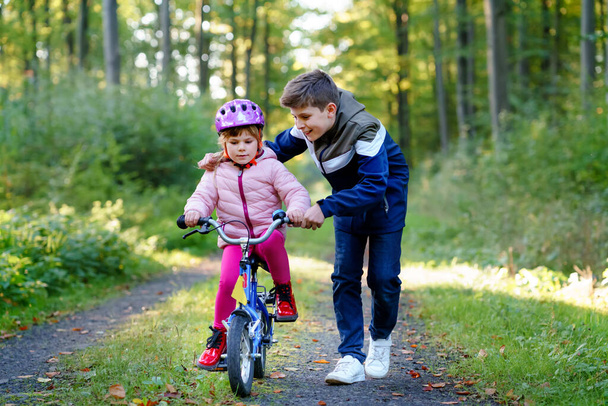 Bisiklet süren güvenlik kaskı takan küçük tatlı bir anaokulu kızı. Okullu çocuk, erkek kardeş mutlu sağlıklı kız kardeş çocuk bisikleti öğretiyor ve bisikletle eğleniyor. Aktif aile fertleri dışarıda. - Fotoğraf, Görsel