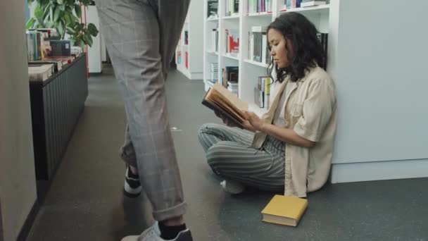 Langzame shot van vrouwelijke Aziatische universiteit student in casualwear zitten op de vloer in de moderne bibliotheek leunend op boekenkast terwijl het lezen van boek - Video
