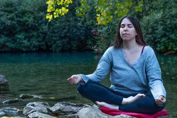 背景に湖がある高い山の川でヨガや瞑想をしている若いブルネットの女の子。青いセーターと黒いパンツを着て - 写真・画像