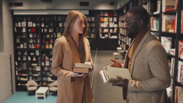 Plan de ralenti moyen de jeunes couples multiethniques en tenue décontractée intelligente debout à la bibliothèque moderne tenant plusieurs livres dans les mains ayant une conversation - Séquence, vidéo