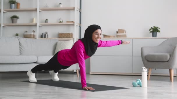 Entrenamiento de fuerza en casa. Joven mujer musulmana activa en hijab practicando ejercicio dinámico de tablón, levantando la mano en el suelo - Metraje, vídeo