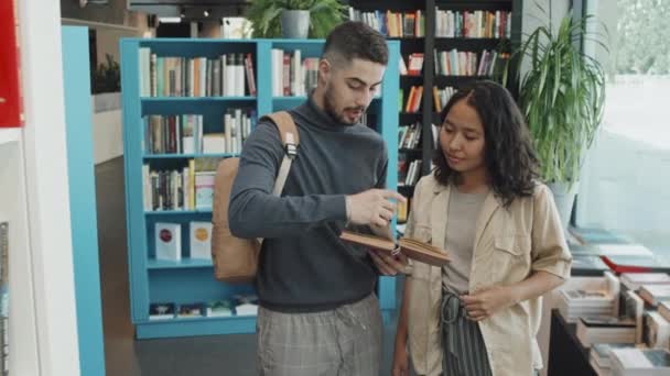 Tiro de lentitud media de dos jóvenes estudiantes universitarios multiétnicos discutiendo el libro mientras están de pie en la biblioteca moderna - Metraje, vídeo