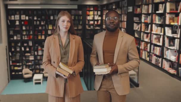 Medium-Slowmo-Porträt zweier junger multiethnischer Universitätsprofessoren in schicker Casualwear, die in der zeitgenössischen Bibliothek mit mehreren Papierbüchern in der Hand für die Kamera posieren - Filmmaterial, Video