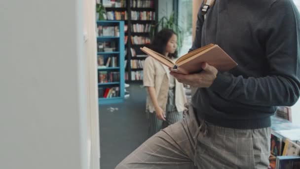 Midsection slowmo shot of unrecognizable male university student standing in front of bookcase in modern library reading book - Footage, Video