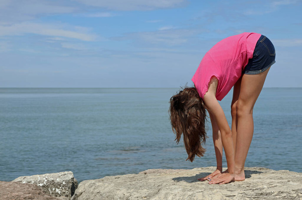 Atletik kız yazın deniz kenarındaki egzersizler sırasında sırtını esnek bir şekilde bükerek ayak parmaklarına dokunur. - Fotoğraf, Görsel