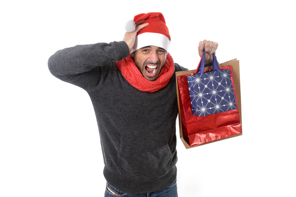  молодий стресовий чоловік у капелюсі Санта тримає червоні сумки для покупок
 - Фото, зображення