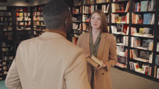 Medium slow shot di giovane donna caucasica con piccolo mucchio di libri in mano che parlano con professore universitario afro-americano in giacca beige in piedi insieme tra le librerie della biblioteca moderna - Filmati, video