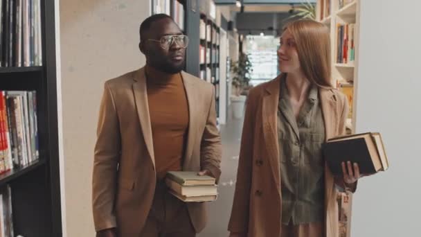 Tracking medium slowmo shot della moderna coppia interrazziale di giovani professori universitari adulti in smart casualwear passeggiando lungo le librerie della biblioteca contemporanea - Filmati, video