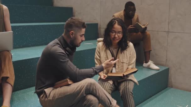 Tiro lento medio de jóvenes estudiantes universitarios multiétnicos sentados en escaleras de color turquesa en interiores estudiando juntos - Imágenes, Vídeo