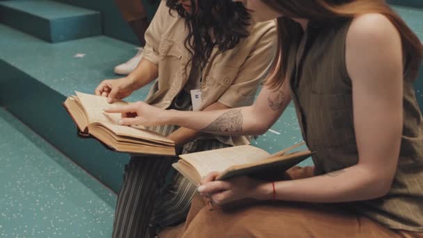 Midsectie slowmo shot van twee onherkenbare vrouwelijke universitaire studenten zittend op turquoise trappen binnen boeken te lezen en de inhoud te bespreken - Video