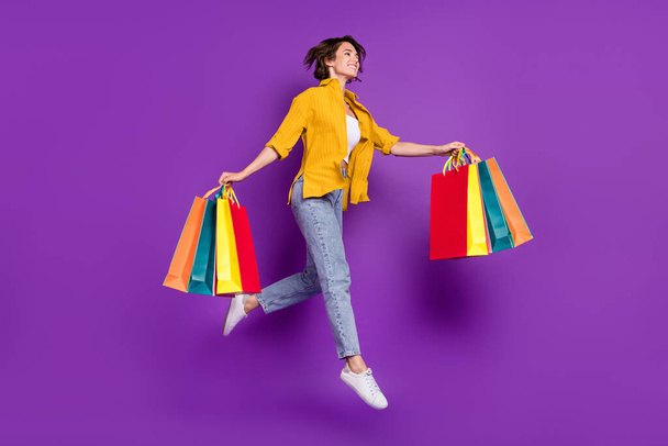 Photo of funky pretty young lady wear yellow shirt jumping high holding shopping bags looking empty space smiling isolated violet color background - Foto, Imagen