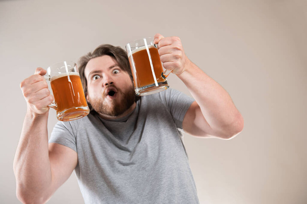 Muotokuva onnellinen nuori parrakas mies tilalla kaksi olutta mukeja eristetty vaaleanharmaa tausta - Valokuva, kuva