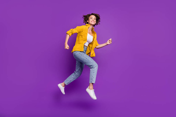 Photo of adorable shiny young woman dressed yellow shirt smiling jumping high running fast isolated purple color background - Photo, image
