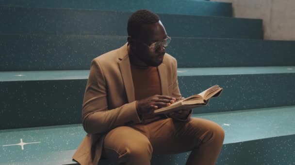 Közepes lassítású felvétel fiatal felnőtt afro-amerikai férfi egyetemi hallgató intelligens alkalmi viselet és szemüveg olvasás papír könyv ül türkiz lépcsőn beltérben - Felvétel, videó