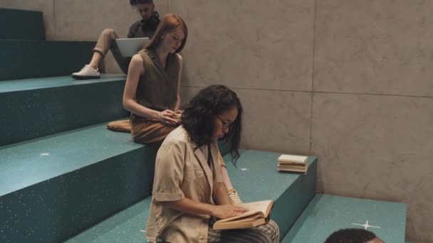 Lassított felvétel fiatal, többnemzetiségű férfi és női egyetemi hallgatókról, akik a lépcsőházban ülnek, egyenként olvasva az újságokat és tanulva - Felvétel, videó