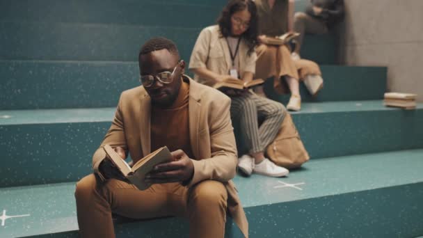 Medium slowmo portrait of African-American young adult university student in smart casualwear and eyeglasses holding paper book and looking at camera sitting on stairs indoors with other students - Footage, Video