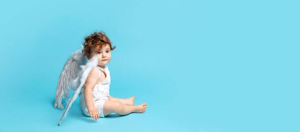 Happy childhood. Little angel. Portrait of cute toddler boy, baby in white romper with angel wings isolated on blue studio background. Concept of childhood, motherhood, life, birth. Copy space for ad - Foto, immagini