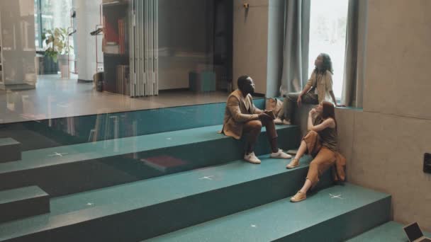 Slowmo ευρύ πλάνο των τριών νέων πολυεθνικών φοιτητών πανεπιστημίου που κάθονται σε τυρκουάζ σκάλες σε εσωτερικούς χώρους έχοντας συνομιλία - Πλάνα, βίντεο