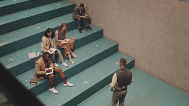 Top-view slowmo shot of group of young adult multi-ethnic university students sitting on turquoise stairs indoors listening to modern male professor during lesson - Footage, Video