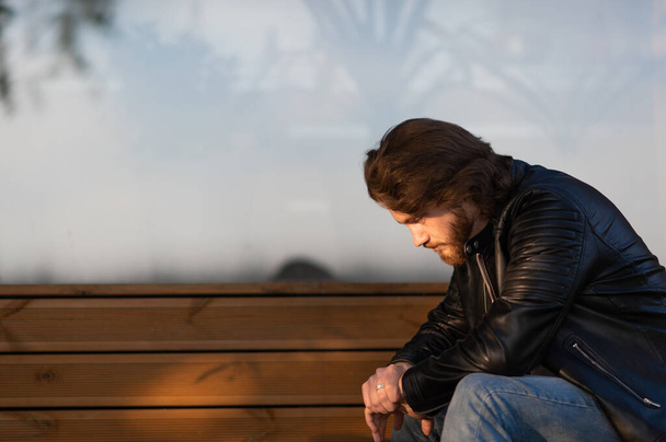 Pensativo hombre barbudo joven serio usar chaqueta de cuero sentado y pensando al aire libre - Foto, Imagen