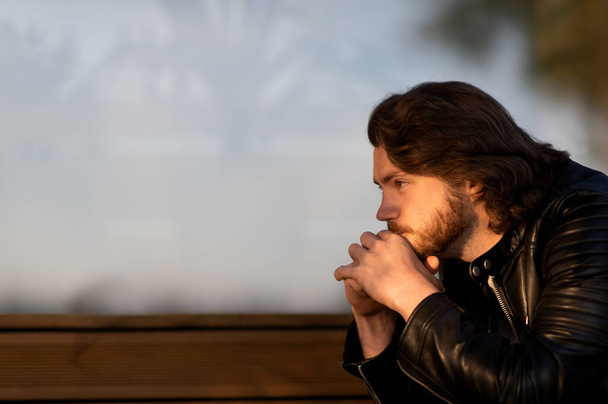 Pensativo hombre barbudo joven serio usar chaqueta de cuero negro sentado y pensando al aire libre - Foto, imagen